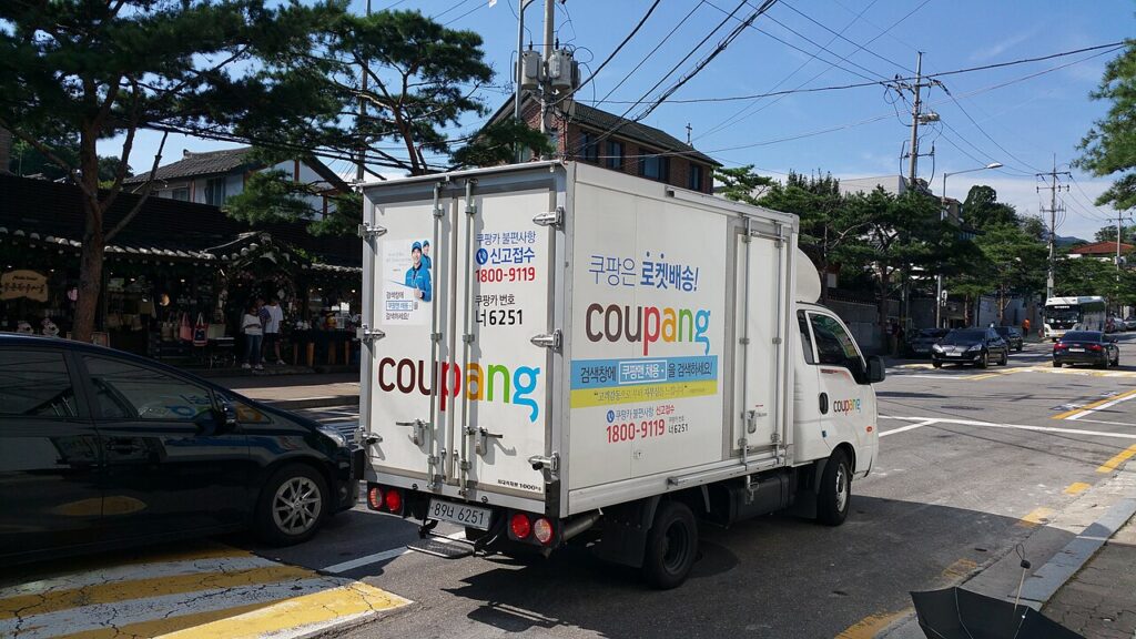 쿠팡 로고와 로켓배송 문구가 적힌 흰색 배송 트럭이 도로 위에 있는 모습